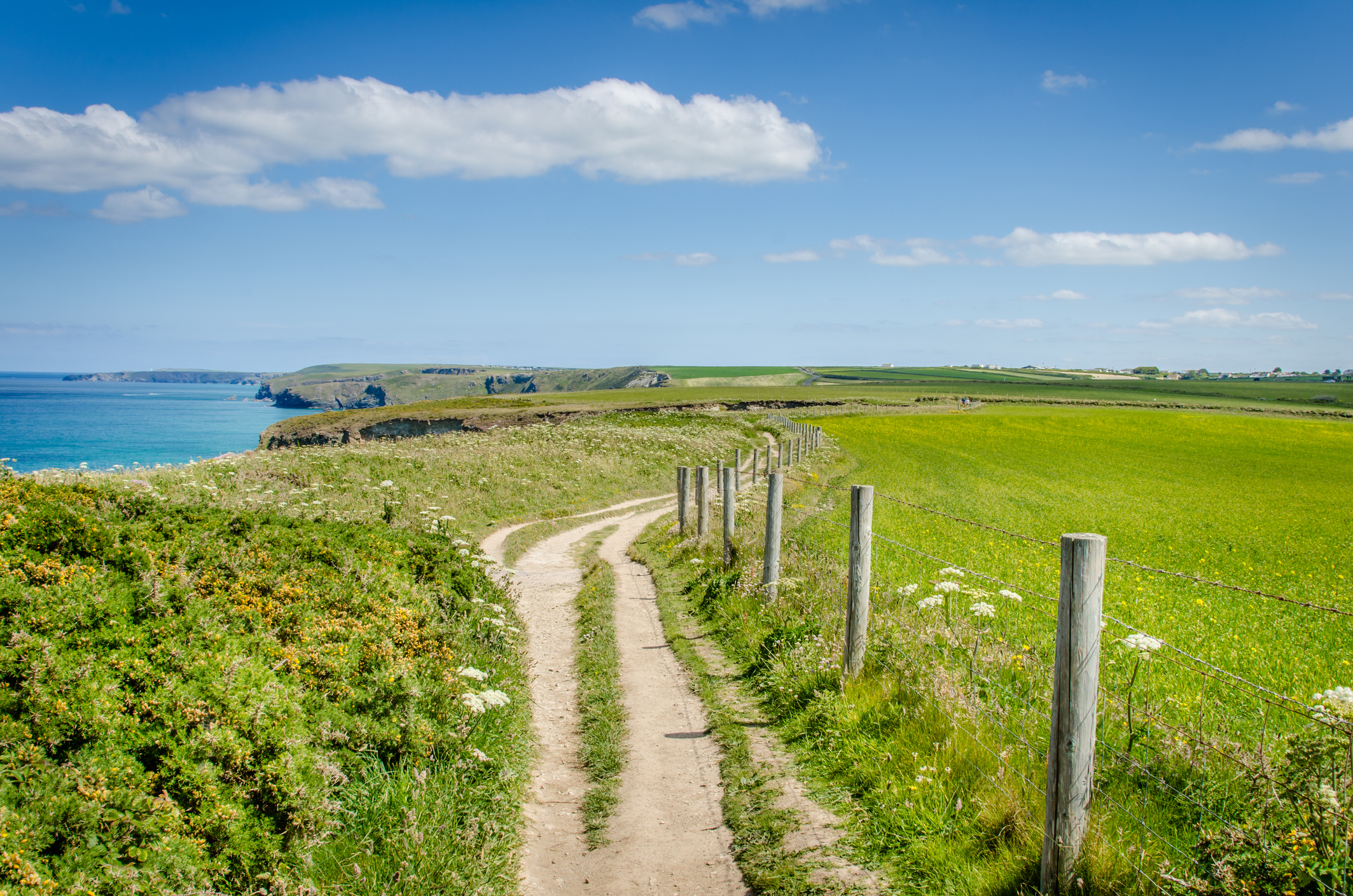 A rural coastal path in Cornwall with a dirt track winding beside green fields and a wooden fence, overlooking the blue sea and cliffs under a bright sky.
