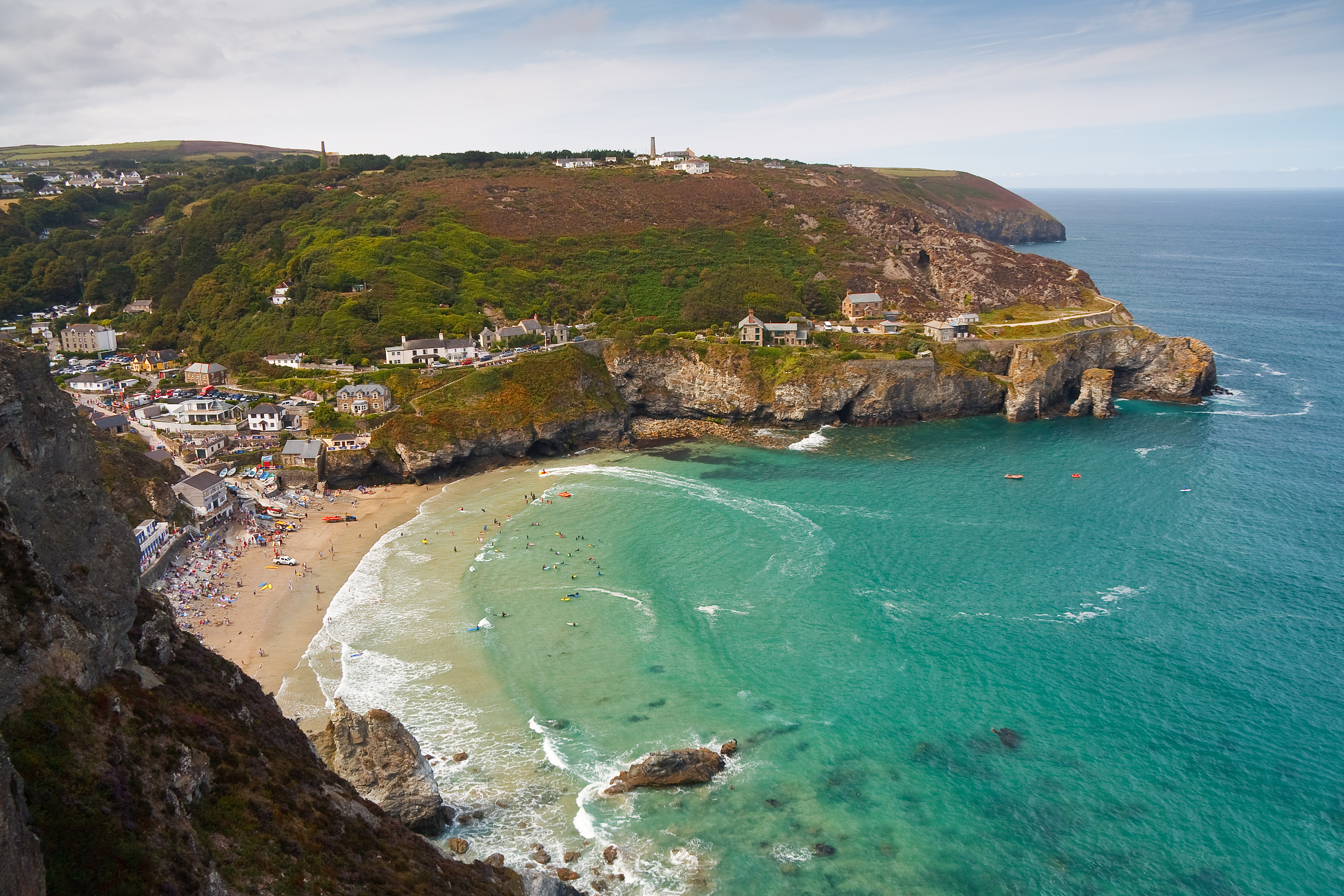 Aerial view of a Cornish seaside village with a sandy cove, cliffs, and turquoise waves with people swimming and relaxing on the beach.