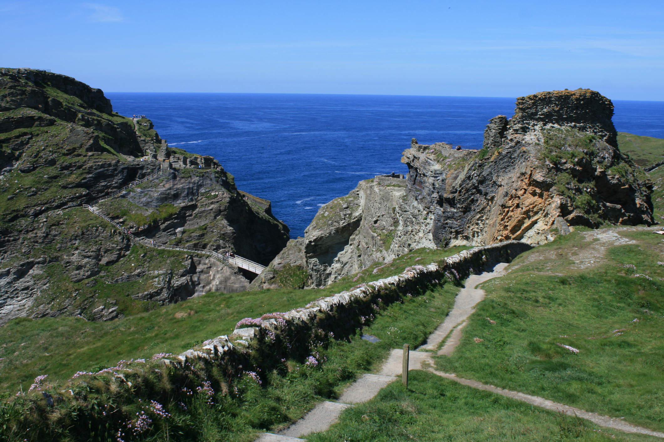 Ruins of Tintagel Castle on dramatic Cornish cliffs with steep rocky paths and wooden bridges overlooking the deep blue Atlantic Ocean.