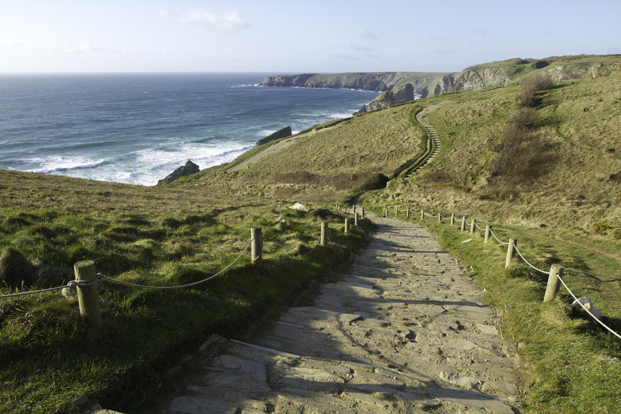 Stone steps descending a grassy Cornish cliff path with wooden posts and rope handrails, leading down towards the sea and coastal headlands.