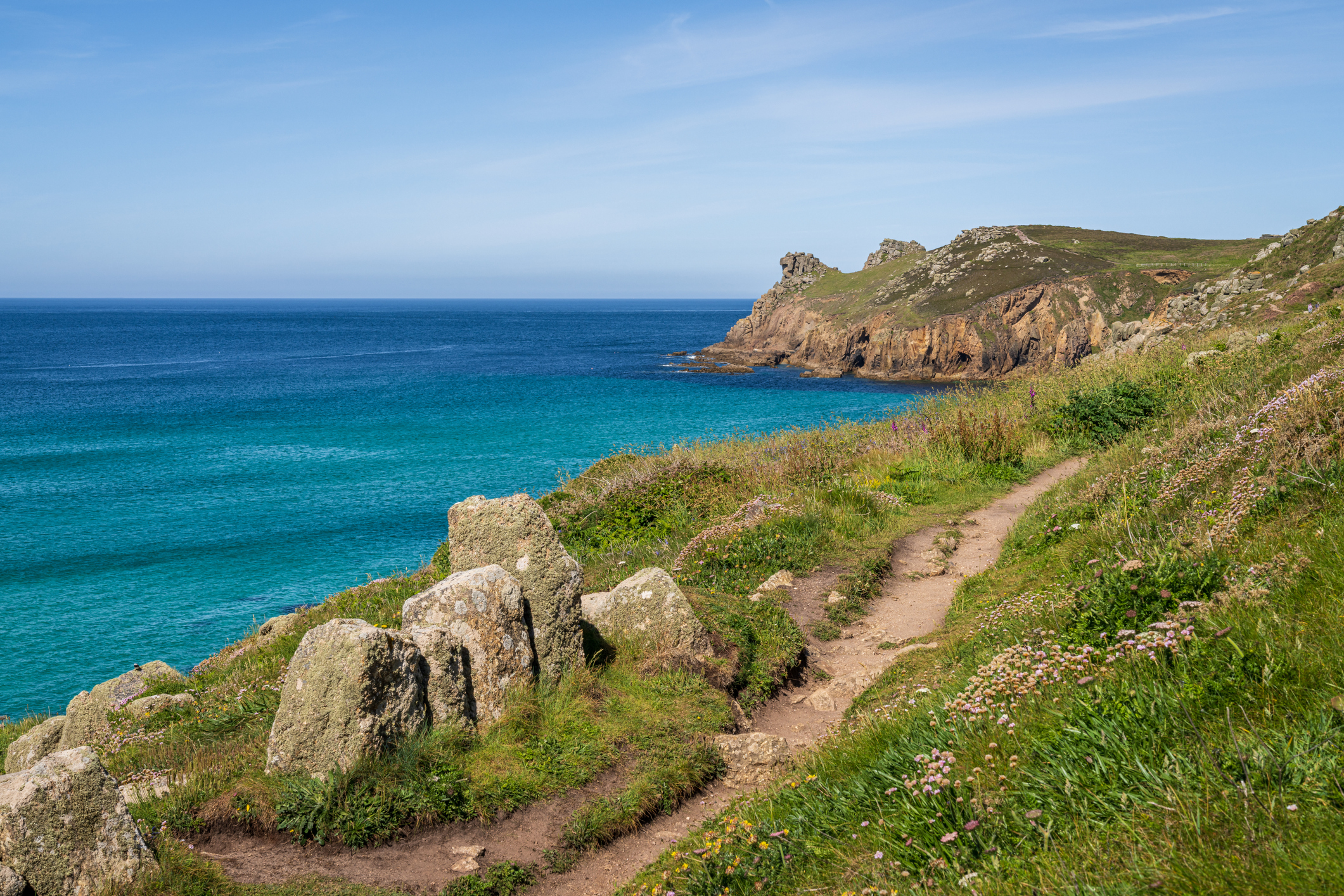 A scenic coastal trail on a clifftop in Cornwall with rugged rocks, grassy slopes, and turquoise sea below under a clear blue sky.